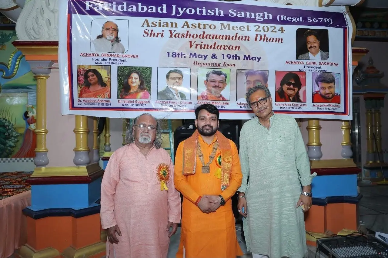 Acharya Tribhuvan Ji with two guests in front of the Asian Astro Meet 2024 banner in Vrindavan