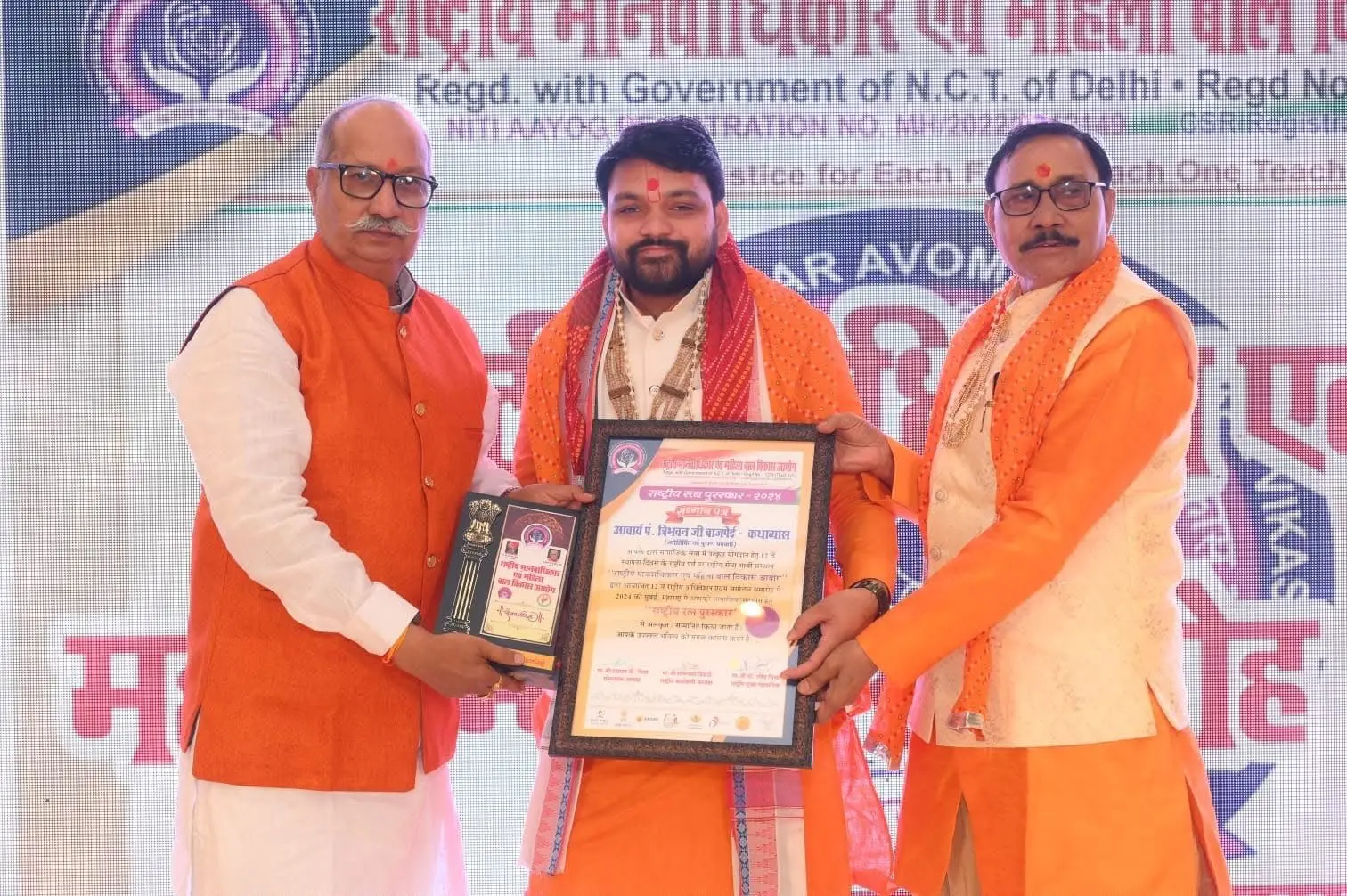 Acharya Tribhuvan Ji receiving the Rashtriya Ratna Puraskar 2024 certificate and memento from two guests
